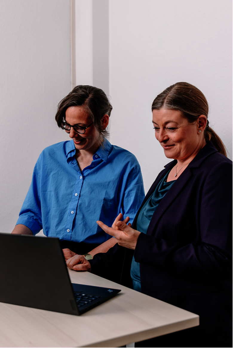 Zwei Frauen im Gespräch an einem Tisch mit einem Computer in einem Büro.